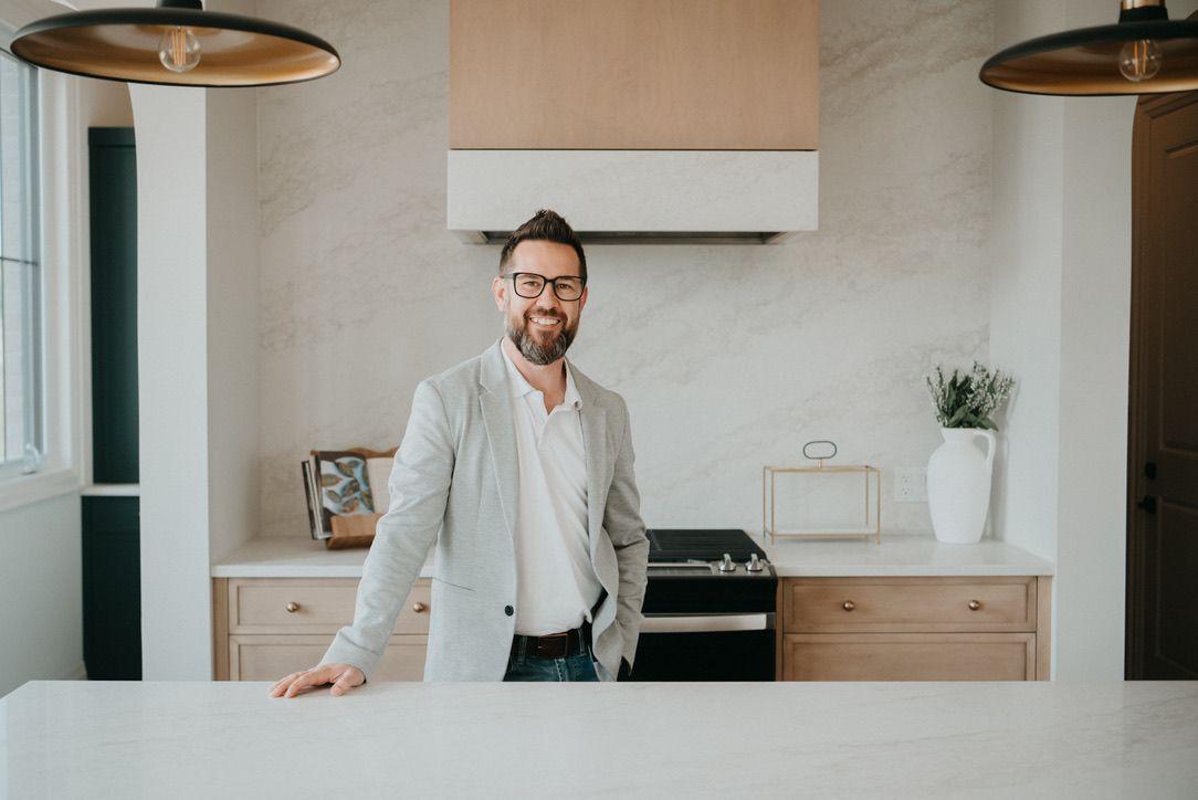 Person standing in a modern kitchen with white countertops and light wooden cabinets, under pendant lights.